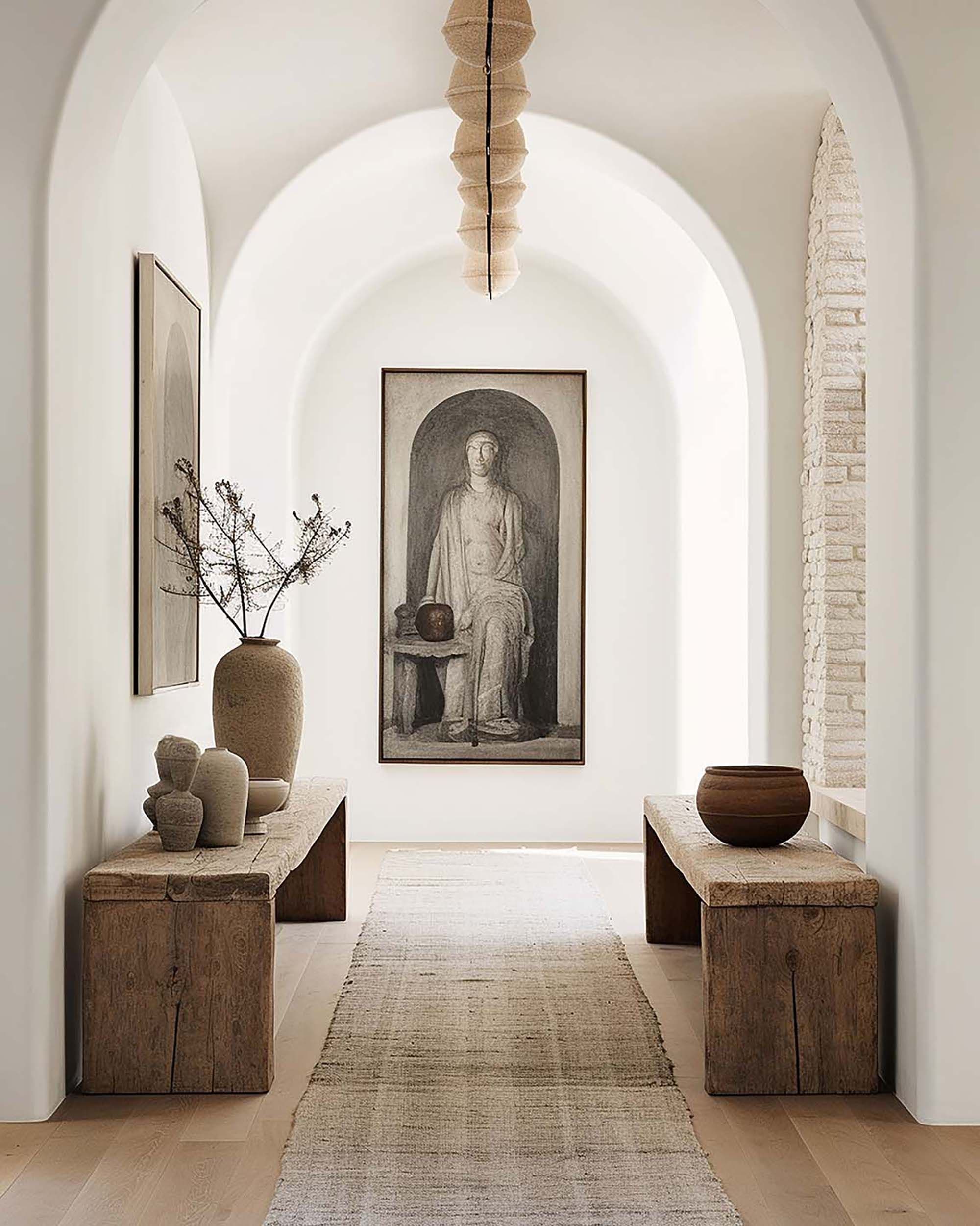 Arched white hallway with a framed artwork on the back wall, flanked by rustic wood benches and a woven pendant light.