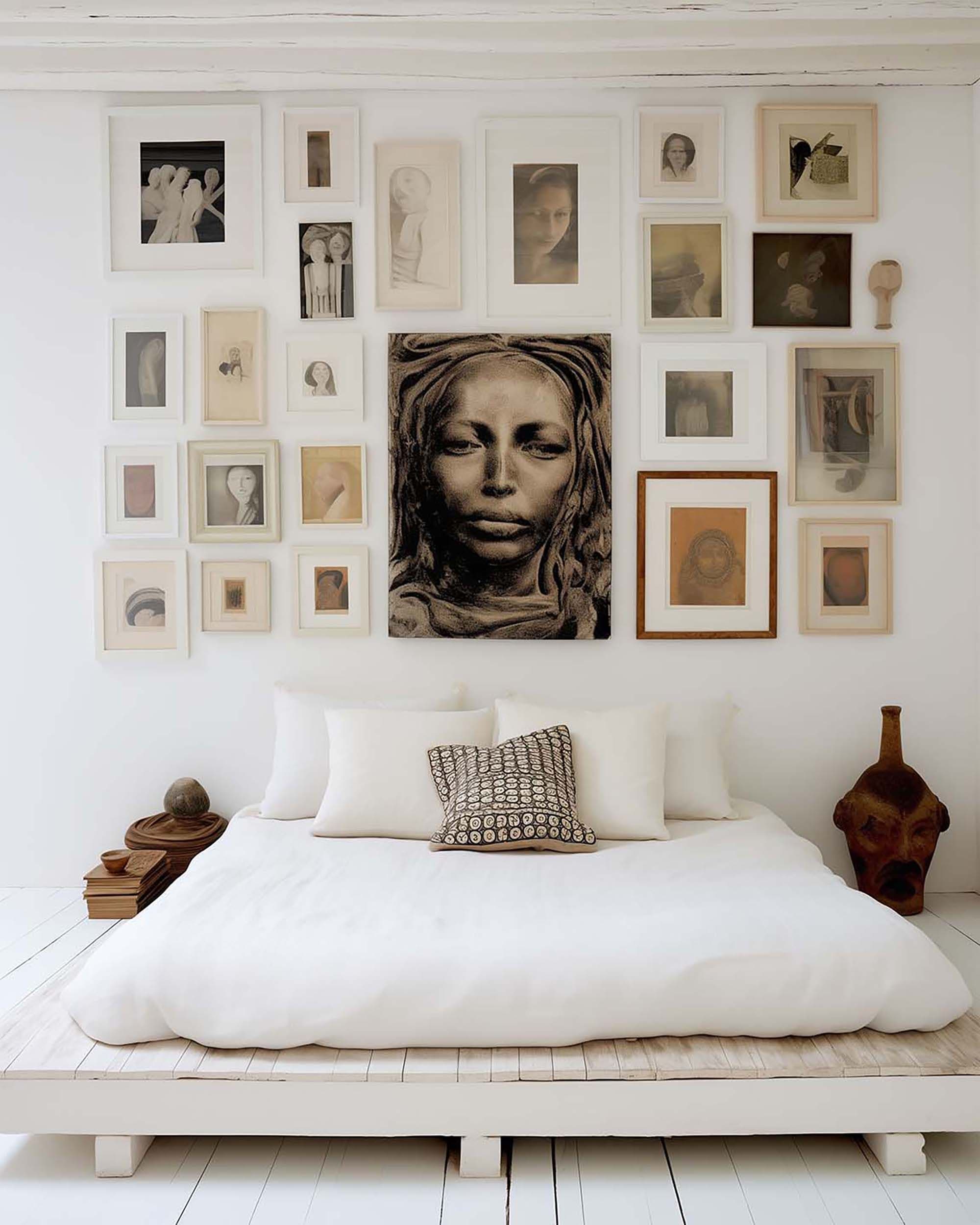 White bedroom with a low platform bed under a gallery wall of assorted frames; wood side table with stacked books beside it.
