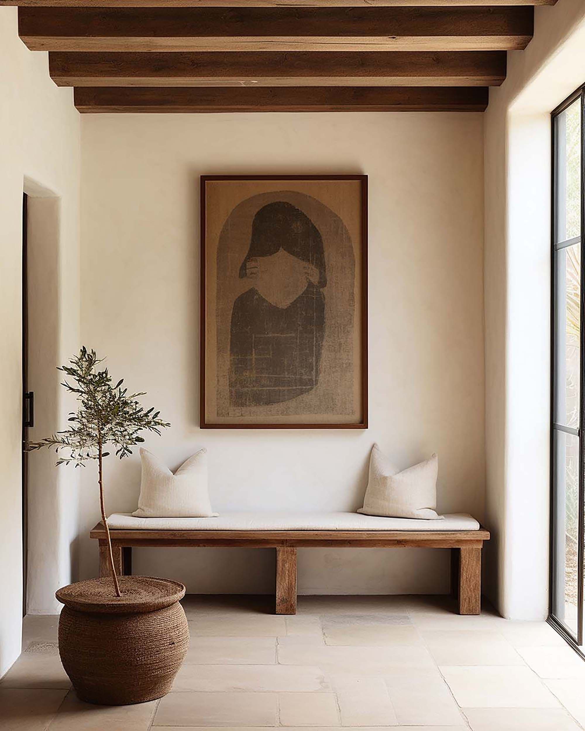 Warm minimalist entryway with a framed artwork above a wood bench with two pillows, beside a tall window.