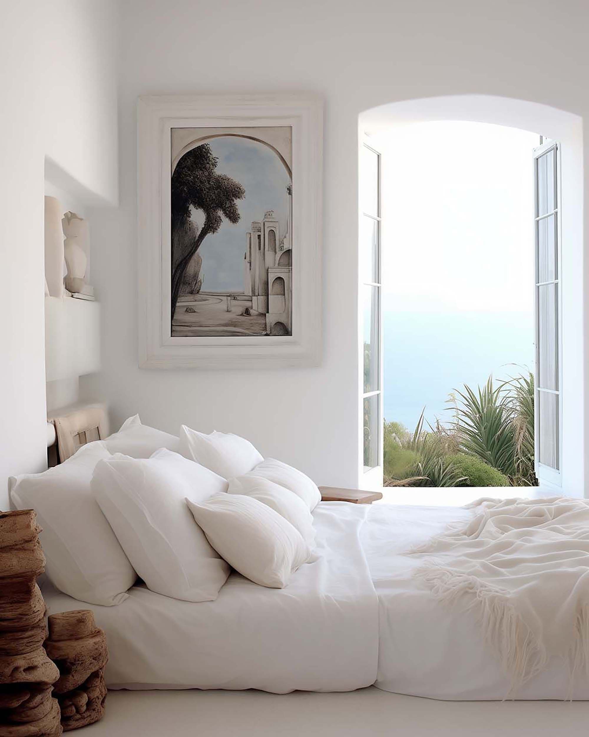 Bright bedroom with a framed wall art piece above a white bed, beside an arched open window with ocean view.