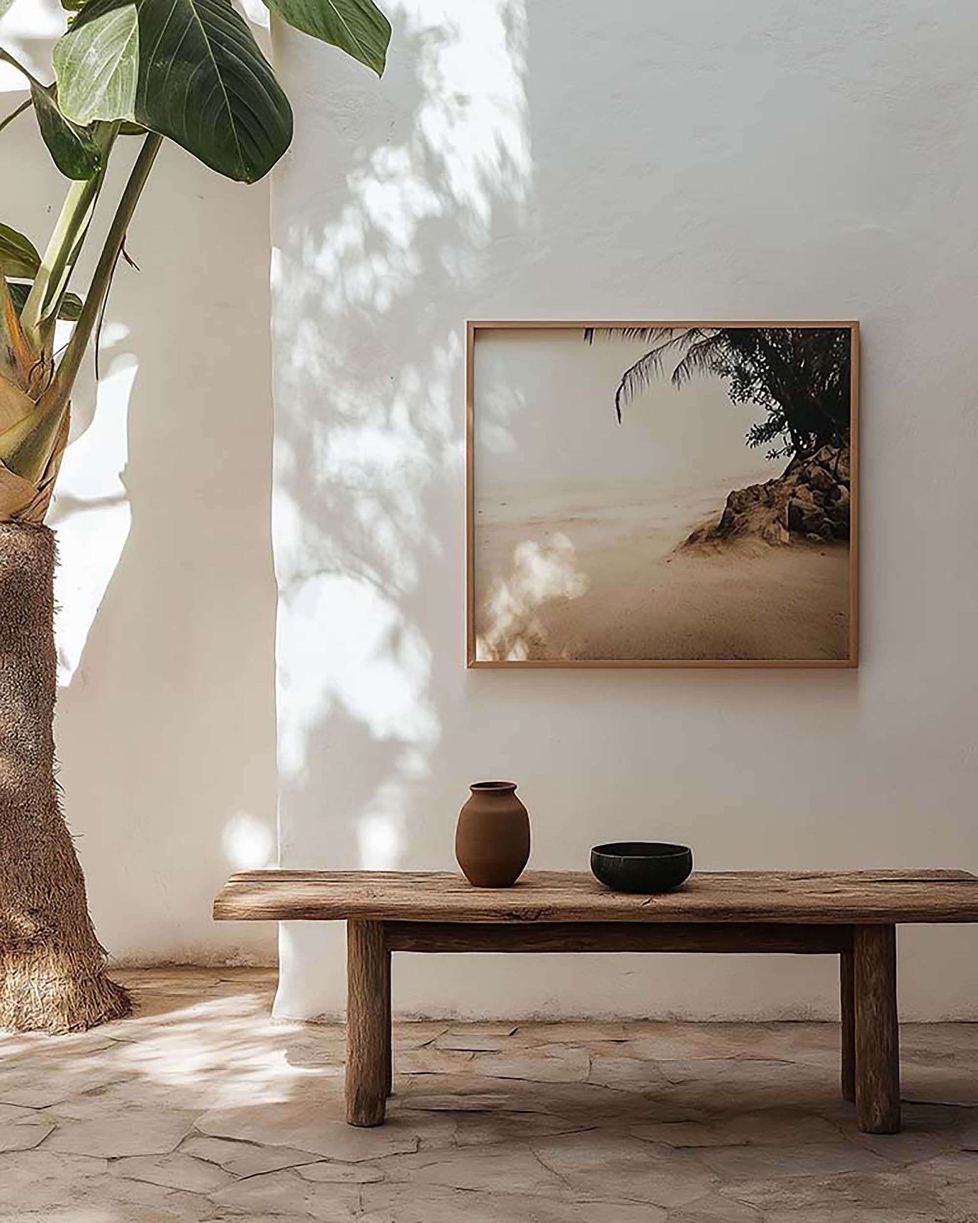 Minimal living room wall with a square wood frame above a rustic wood bench, with soft daylight shadows and a large plant at left.