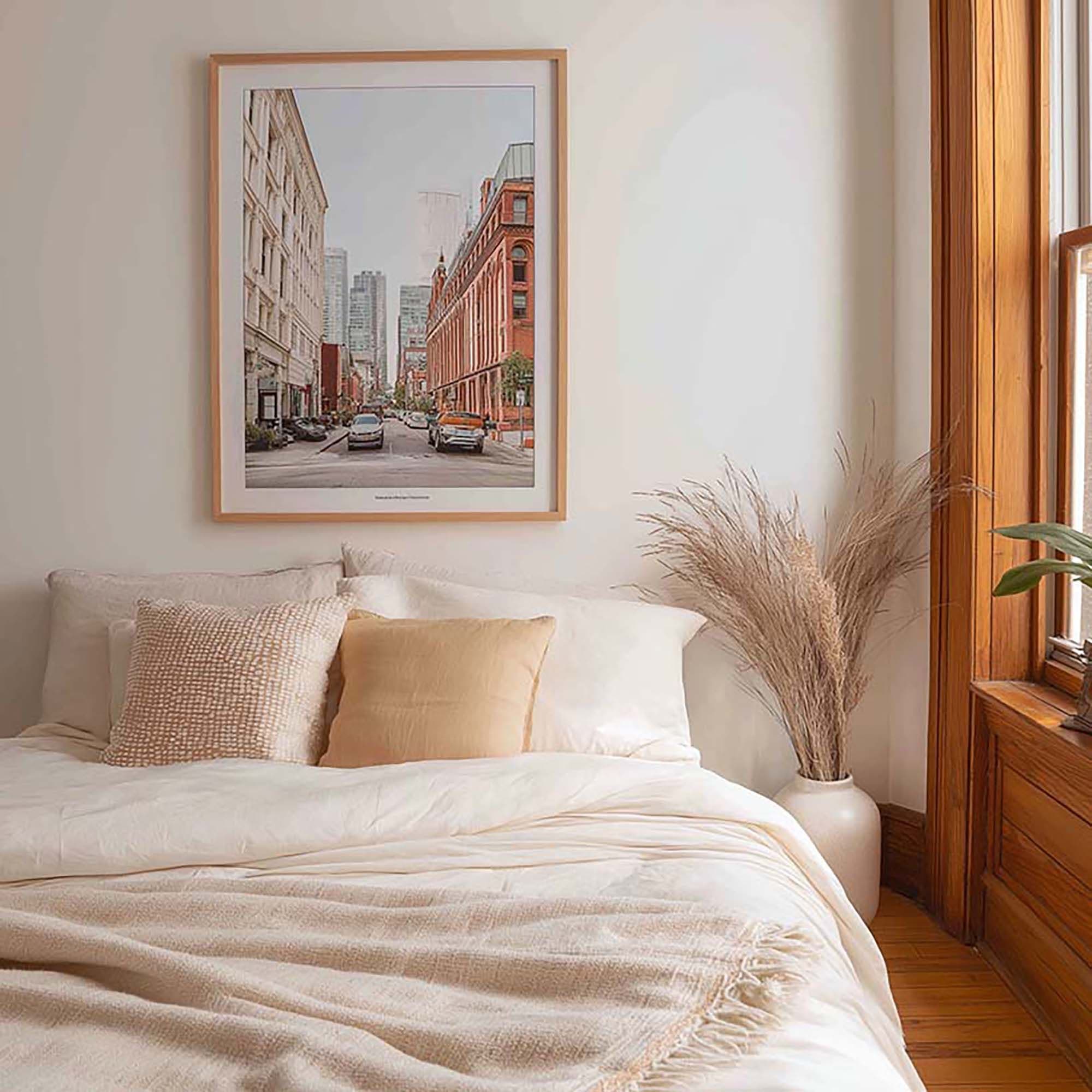 Sunlit bedroom with a framed wall print centered above a made bed, next to a tall vase of dried grass by a wood-trim window.