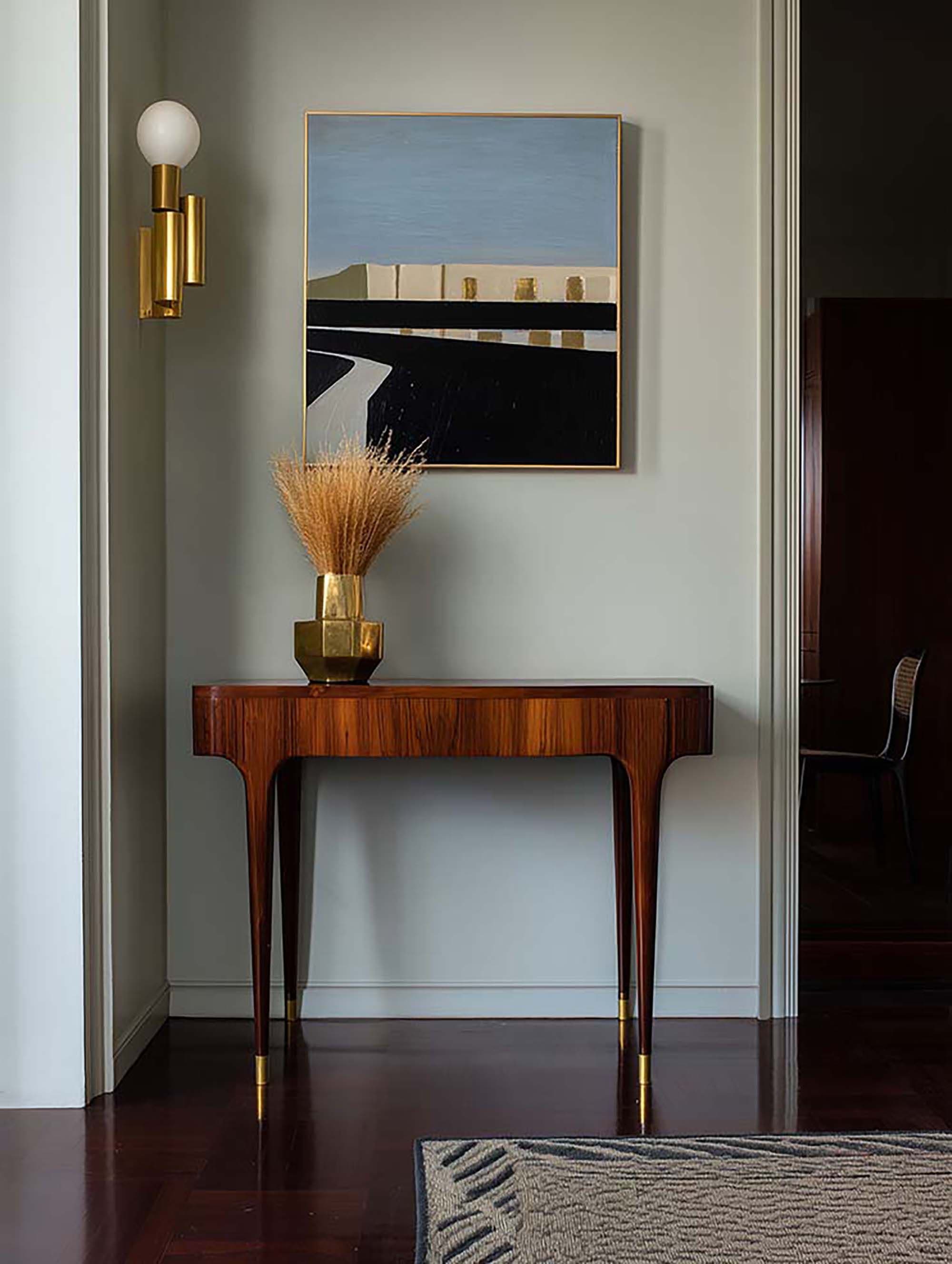 Entryway wall with a framed artwork above a wood console table, with a brass sconce and a vase of dried stems.