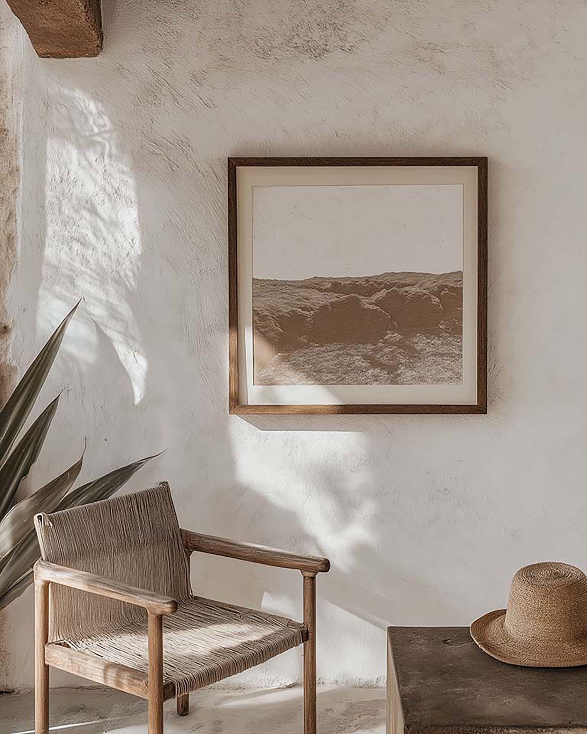 Sunlit living room wall with a square wood frame on textured plaster above a woven lounge chair and a low table.
