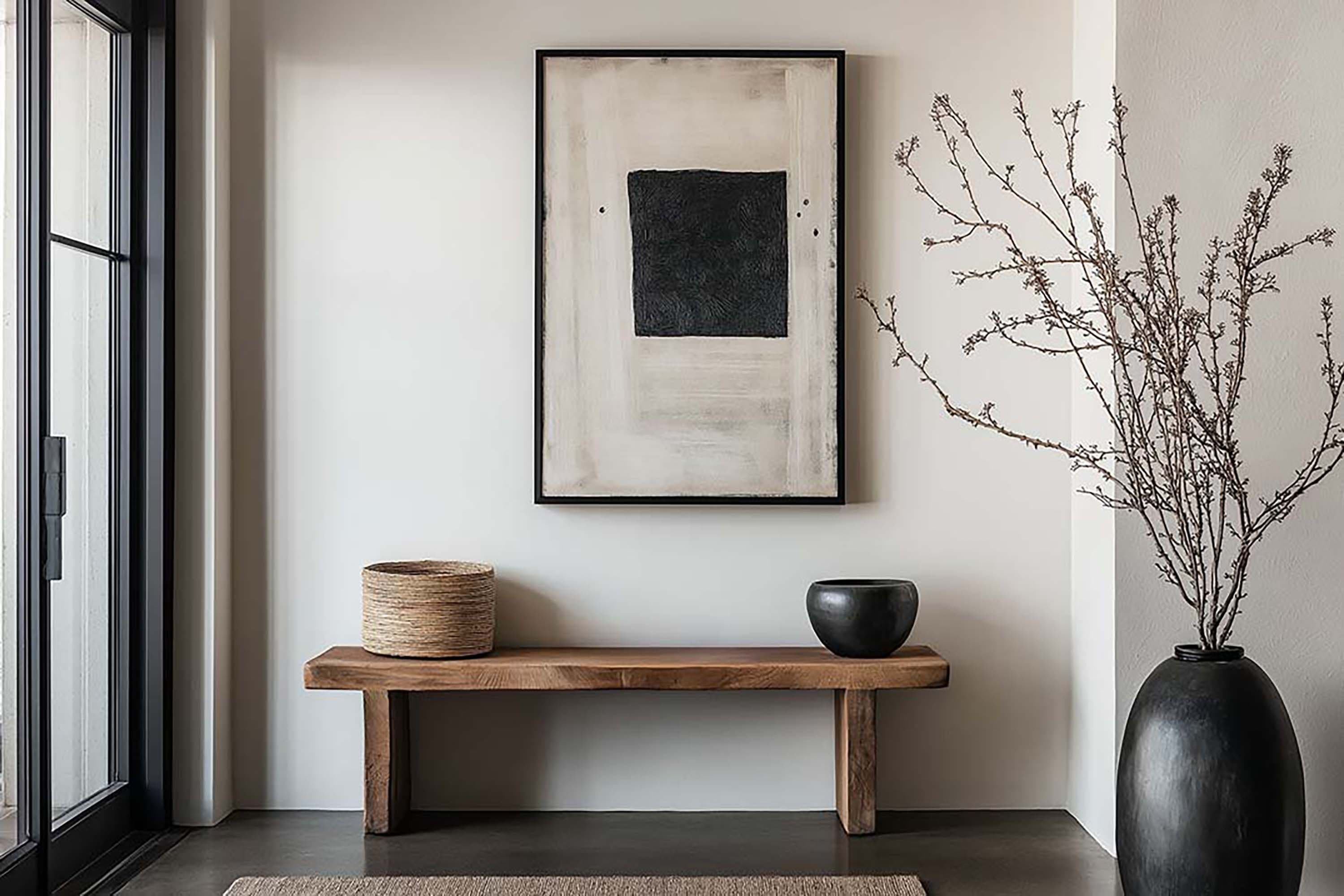 Minimal entryway wall with a black-framed artwork above a low wooden bench, beside a tall black vase with branches.