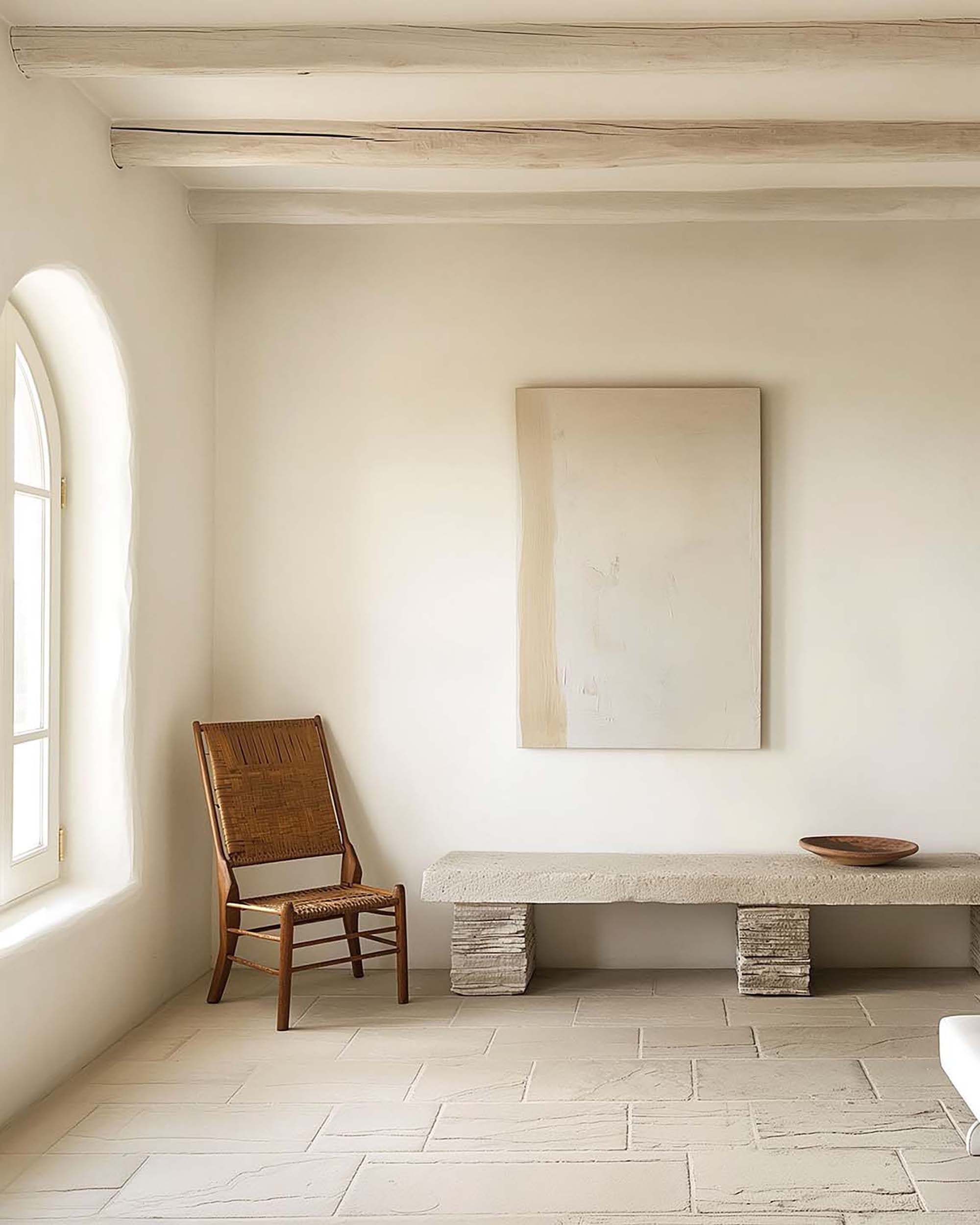Minimal living room with a customizable wall frame above a stone bench, beside a woven chair and arched window.