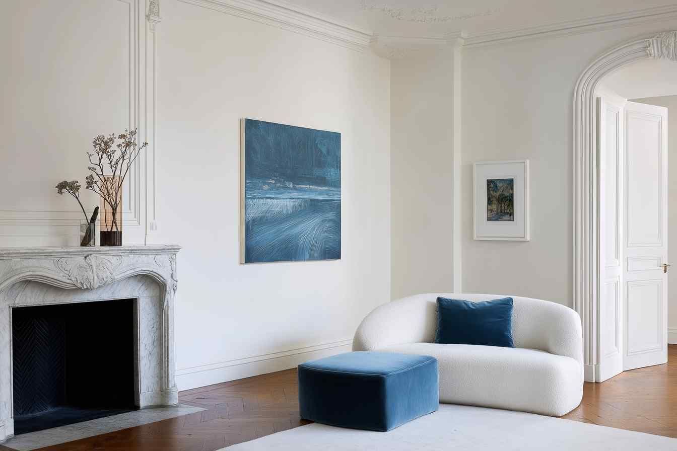 Bright living room with framed wall art above a white curved sofa, next to an ornate marble fireplace and blue ottoman.