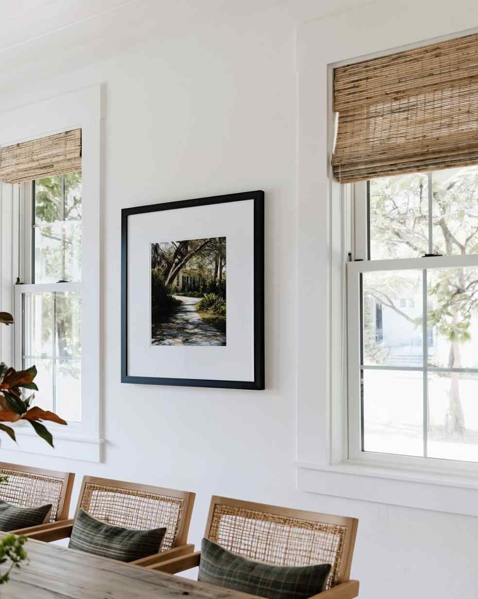 Bright dining room wall with a black framed print between two windows with woven bamboo shades above cane-back chairs.
