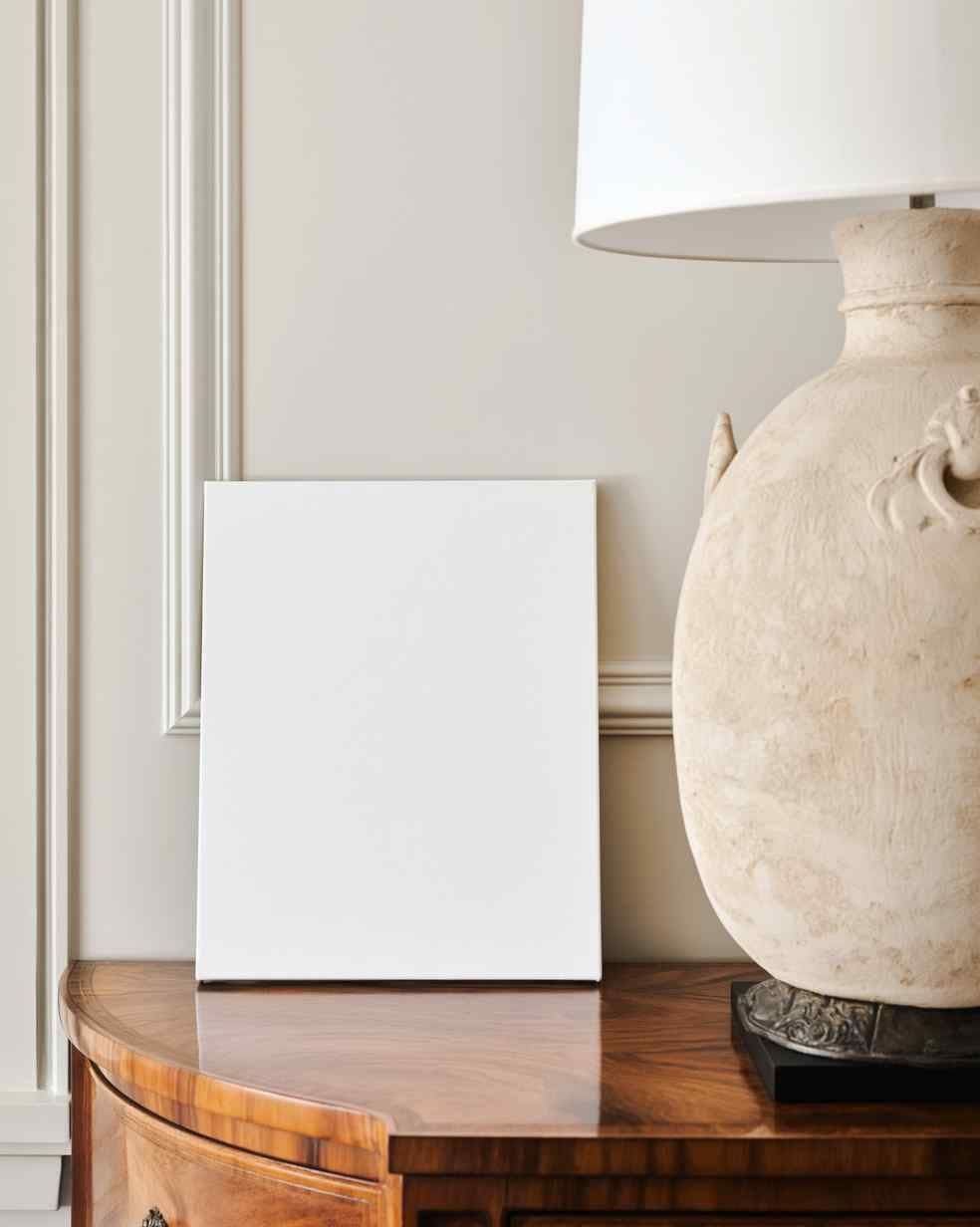 Leaning white frame on a warm wood console table beside a large ceramic table lamp in a traditional entryway.
