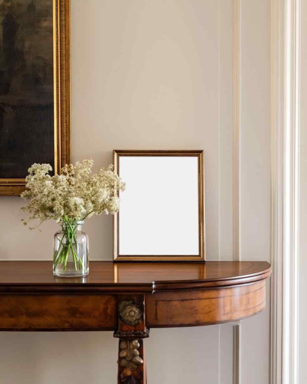 Entryway console table with a gold frame leaning against a light wall, next to a glass vase of flowers.