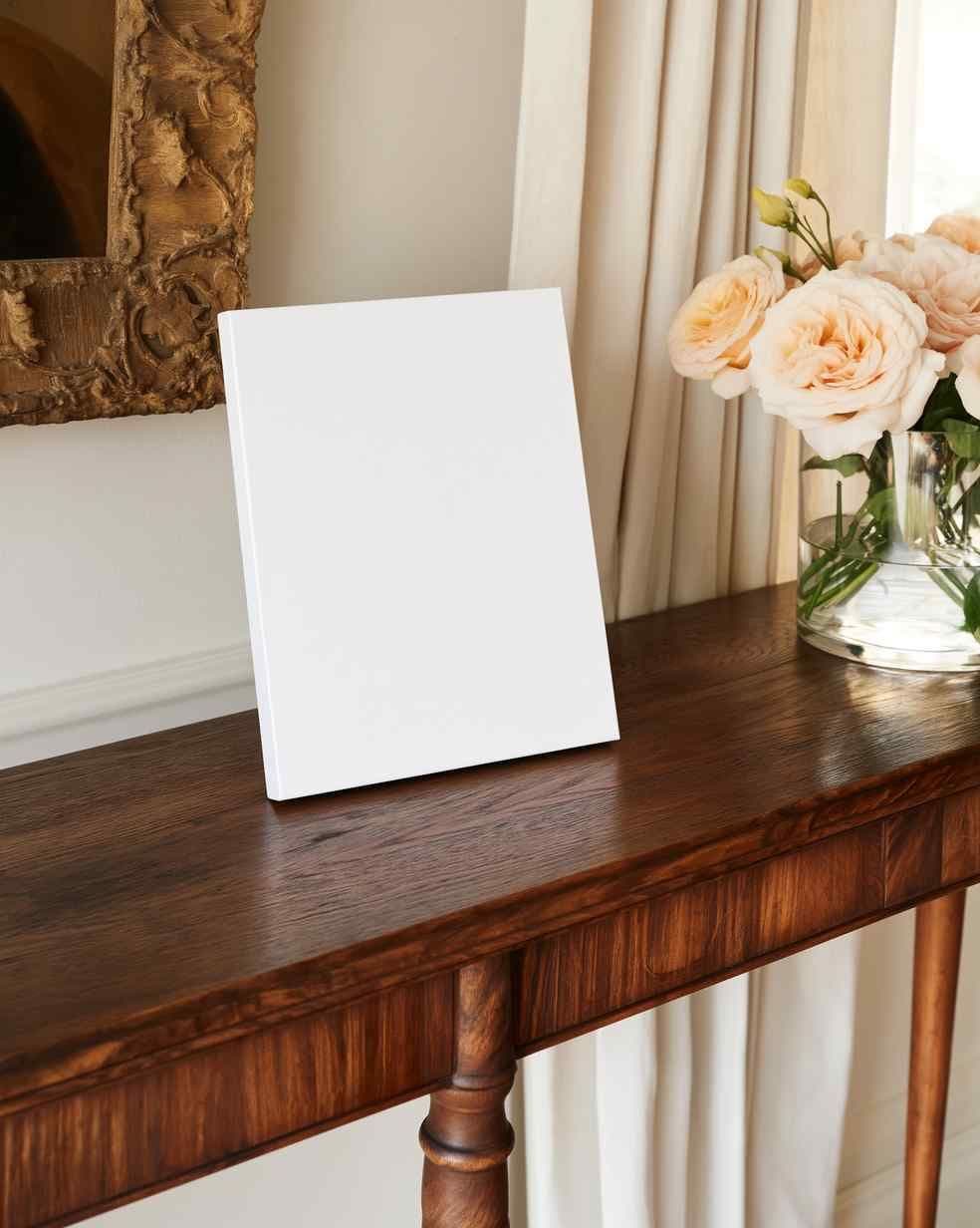 Leaning blank canvas on a dark wood console table beside a glass vase of roses and beige curtains in a living room.