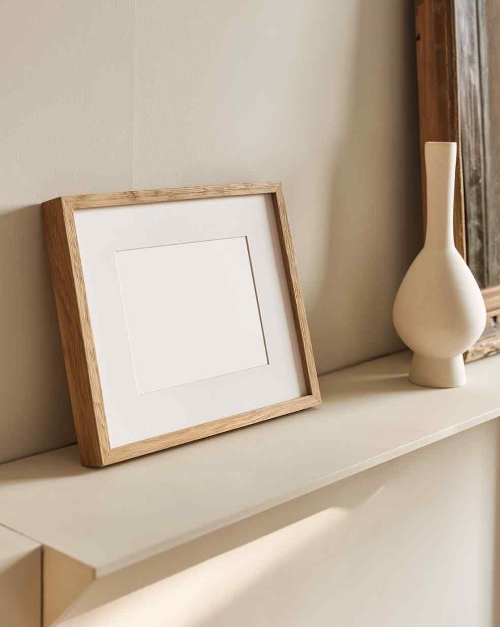 Leaning light-wood frame on a cream console against a beige wall, next to a white ceramic vase near a mirror.