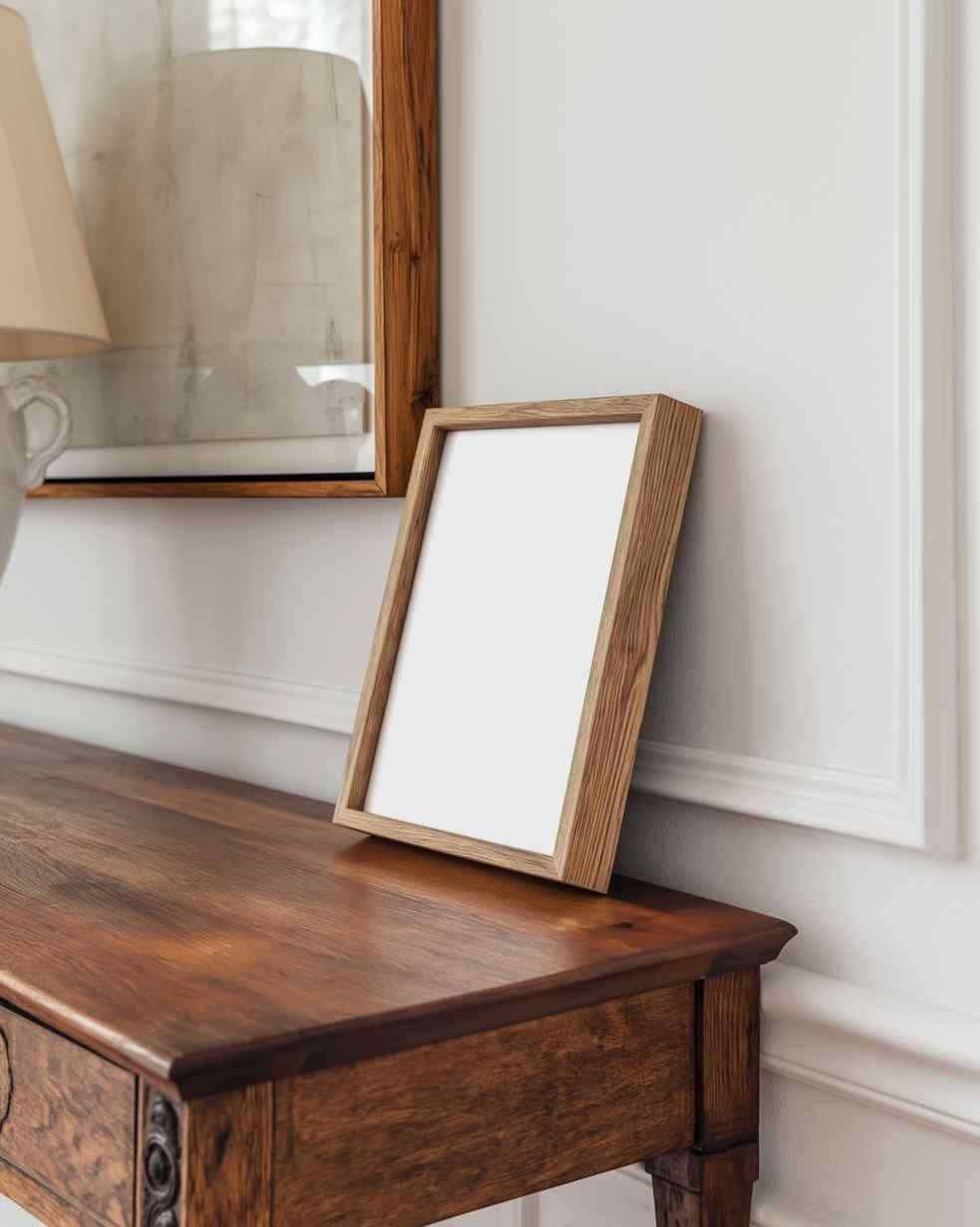 Leaning wood frame on a dark wood console table against a white wall with trim, with a partial wall mirror and lamp visible.