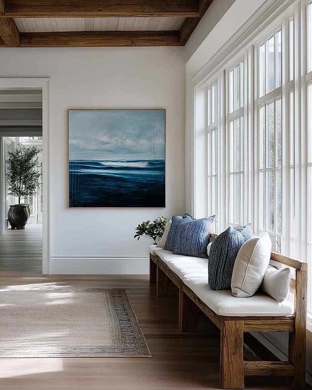 Bright entryway with a framed wall artwork above a wooden bench with pillows, beside tall grid windows.