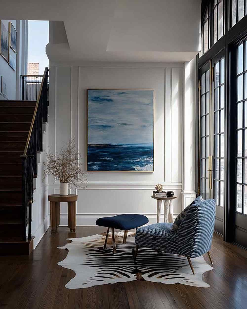 Bright entryway wall with a large framed artwork between white molding, beside a blue chair and tall grid windows.