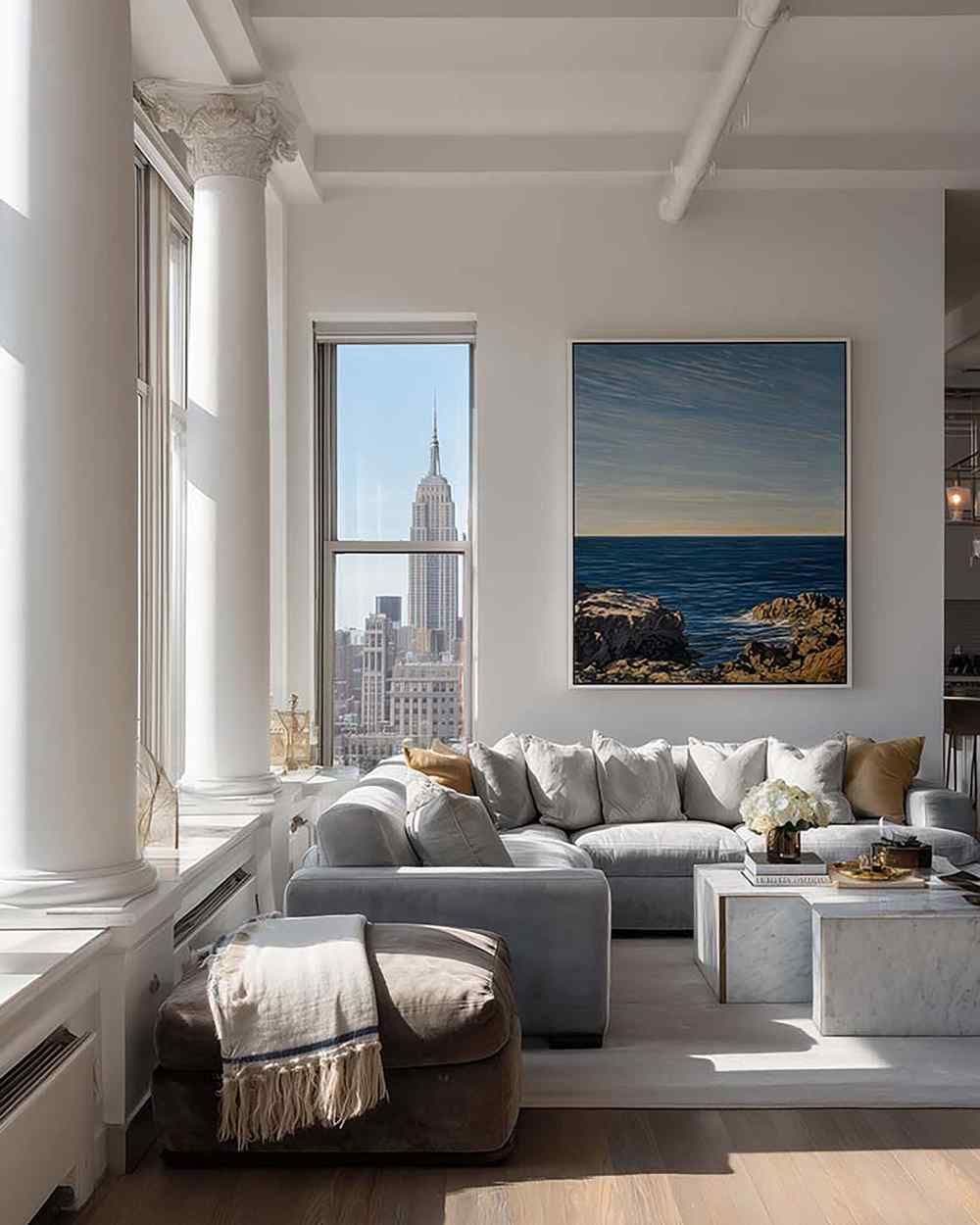 Bright loft living room with a large framed wall art above a gray sectional sofa and a marble coffee table by tall windows.