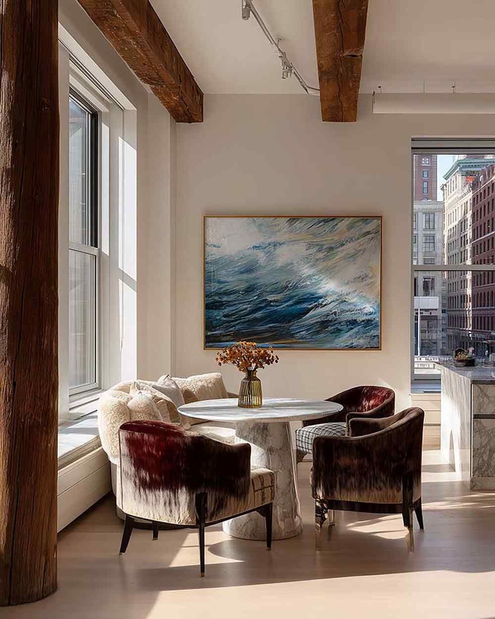 Sunlit dining room with a large framed artwork on a white wall above a round marble table and upholstered chairs.